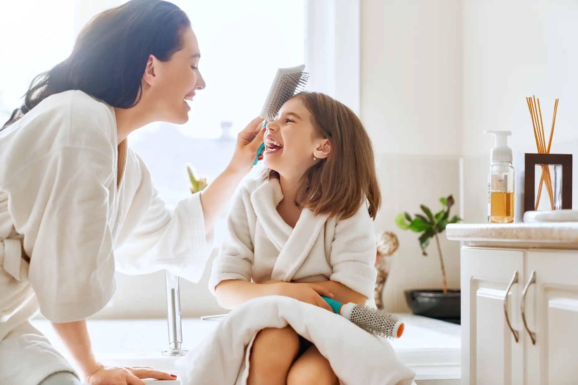 A mom and daughter in the bathroom brushing their hair and laughing.