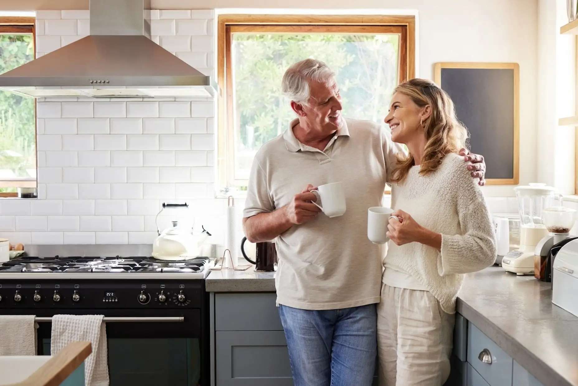 A woman and a man standing in the kitchen smiling, drinking coffee.