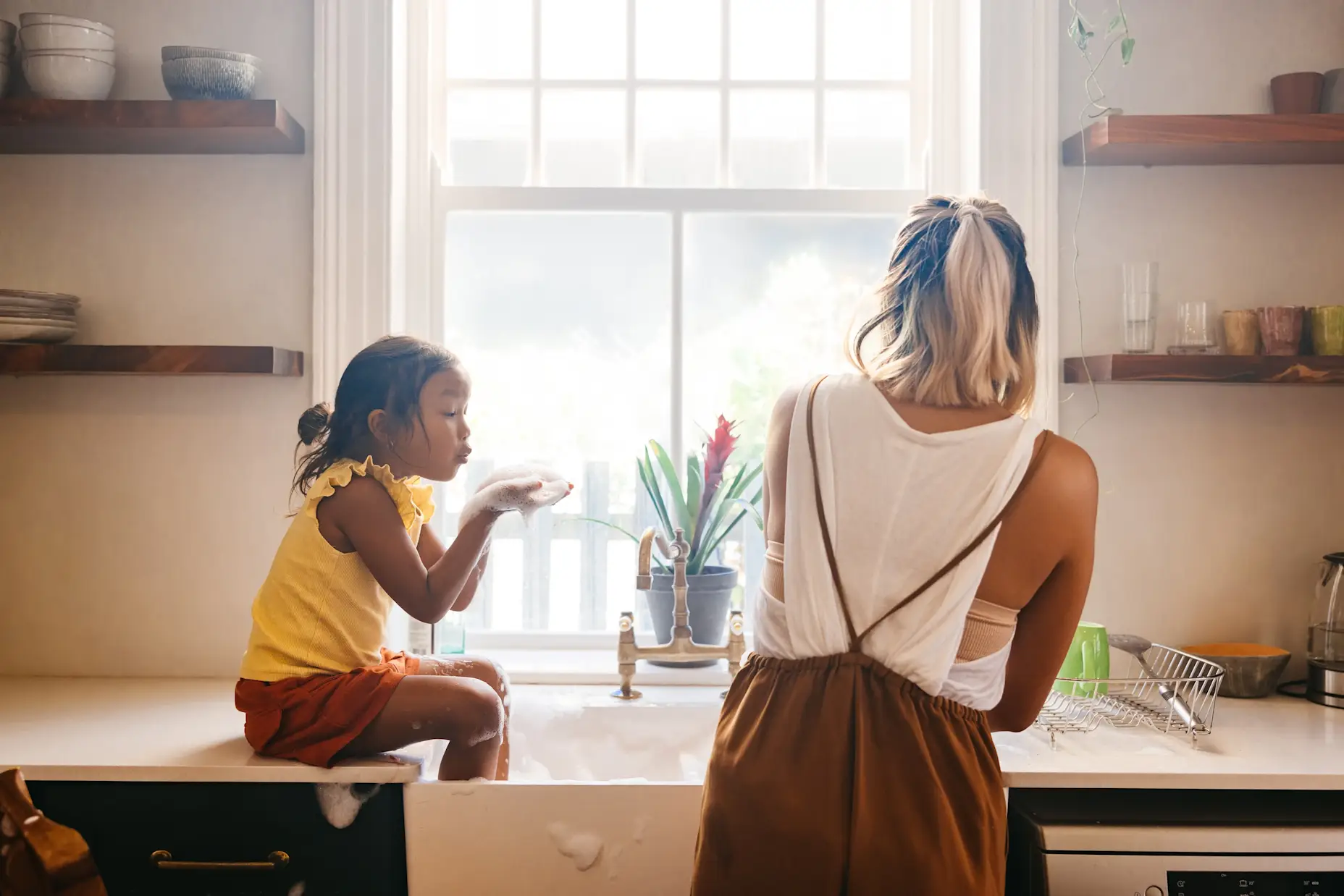 A woman and her daughter blowing bubbles in the sink in front of a window.