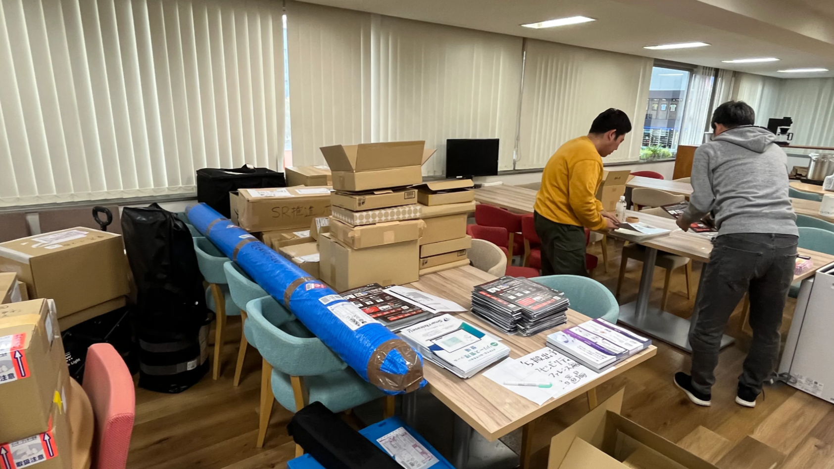 写真：ダンボールや機材で手前のテーブルが埋まっている様子。奥のテーブルで社員2名がチラシの封入作業をしている。