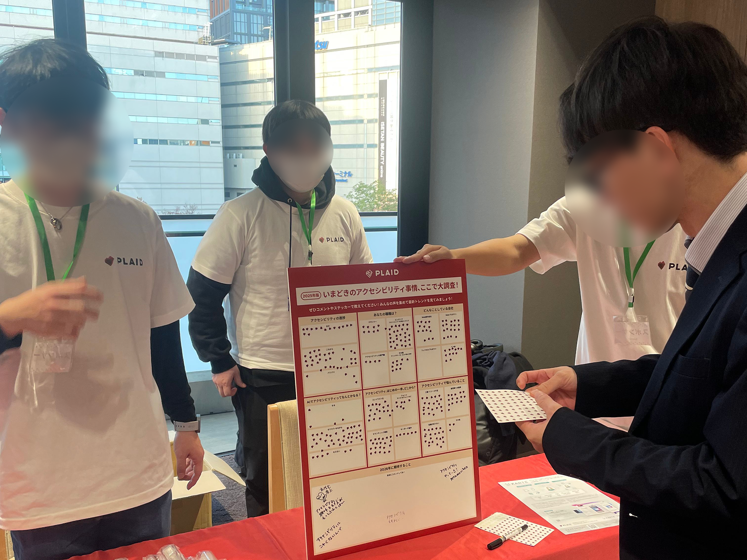 写真：PLAIDさんのブース。テーブルに赤い生地に白字で「PLAID」と書かれたテーブルクロスがかけられ、パネルにシールを貼る形式でアンケートをとっている。ノベルティを配布している。