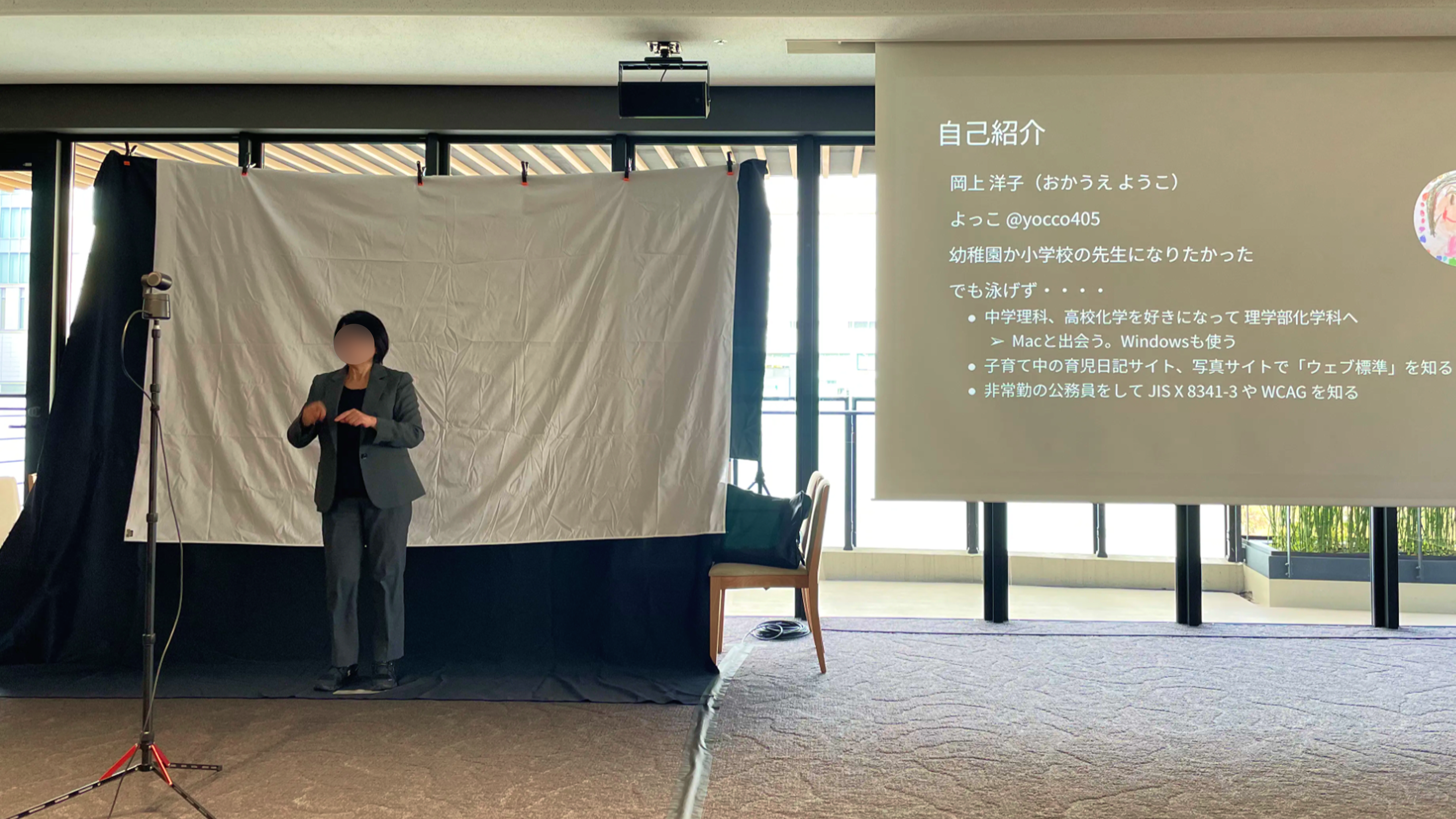 写真：スクリーンを見て左手に設置した手話通訳者による同時通訳の様子。手話通訳者の後ろに暗幕が張られている。