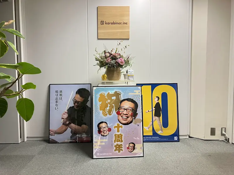 オフィスのエントランスの写真。社名の看板が壁に掛けられ、受付台に花が飾られている。受付台の周りに10周年を記念したパネルポスターが3種類掲示されている。