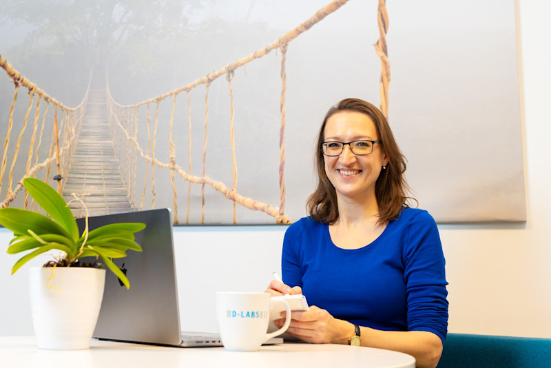 Eine Frau, die hinter einem Computerbildschirm sitzt und Notizen macht. Sie schaut in die Kamera.