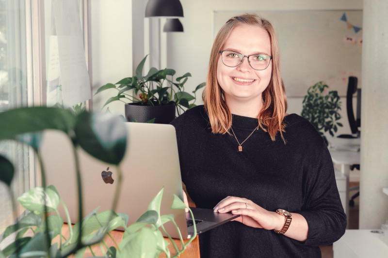Teammitglied Stella sitzt lächelnd vor ihrem Laptop im Berliner Büro. Im Hintergrund sind Pflanzen zu sehen.