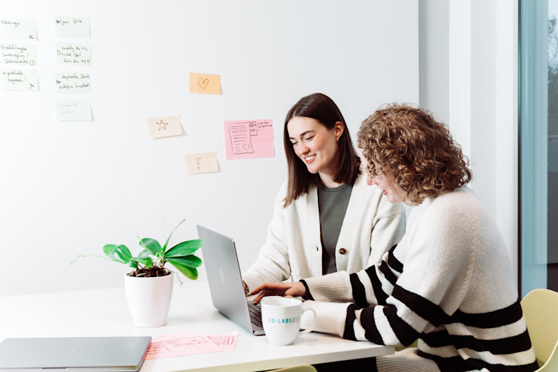 Zwei User Researcher sitzen gemeinsam am Laptop und sprechen. Im Hintergrund sind Post-its auf einem Whiteboard zu sehen.