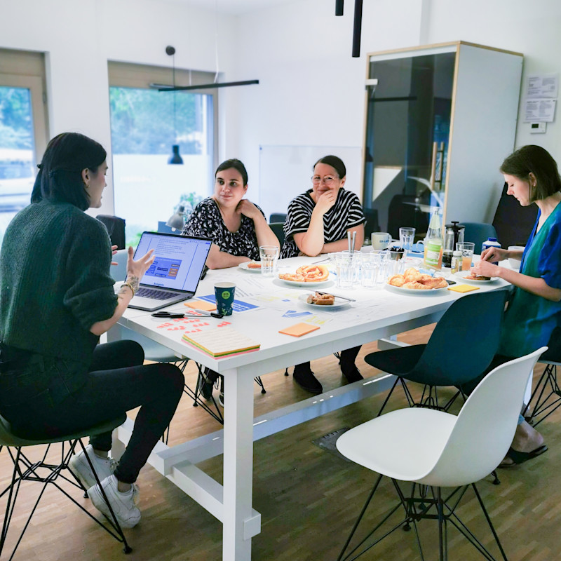 A busy working session with pretzels, croissants and drinks. Four people sit at a table in a bright office space, engrossed in conversation.