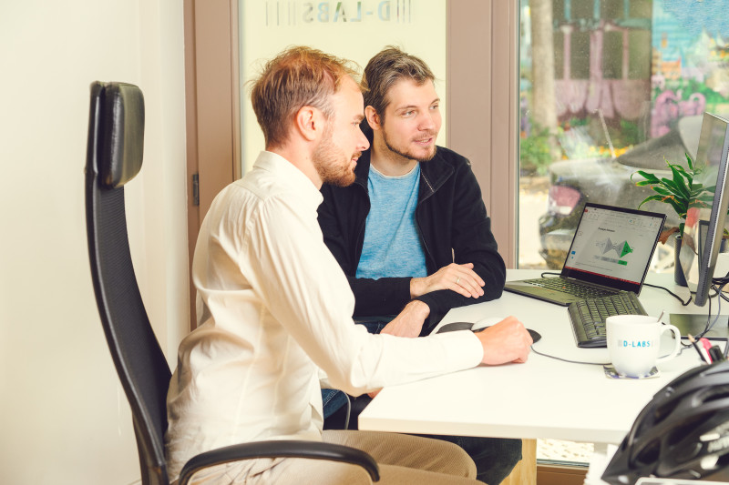 Two men sit in front of a laptop and work together on a project.