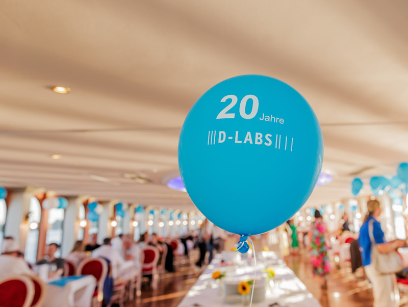 A blue party balloon reading 20 Years of D-LABS. In the background the inside of the MS Sanssouci, decorated blue.