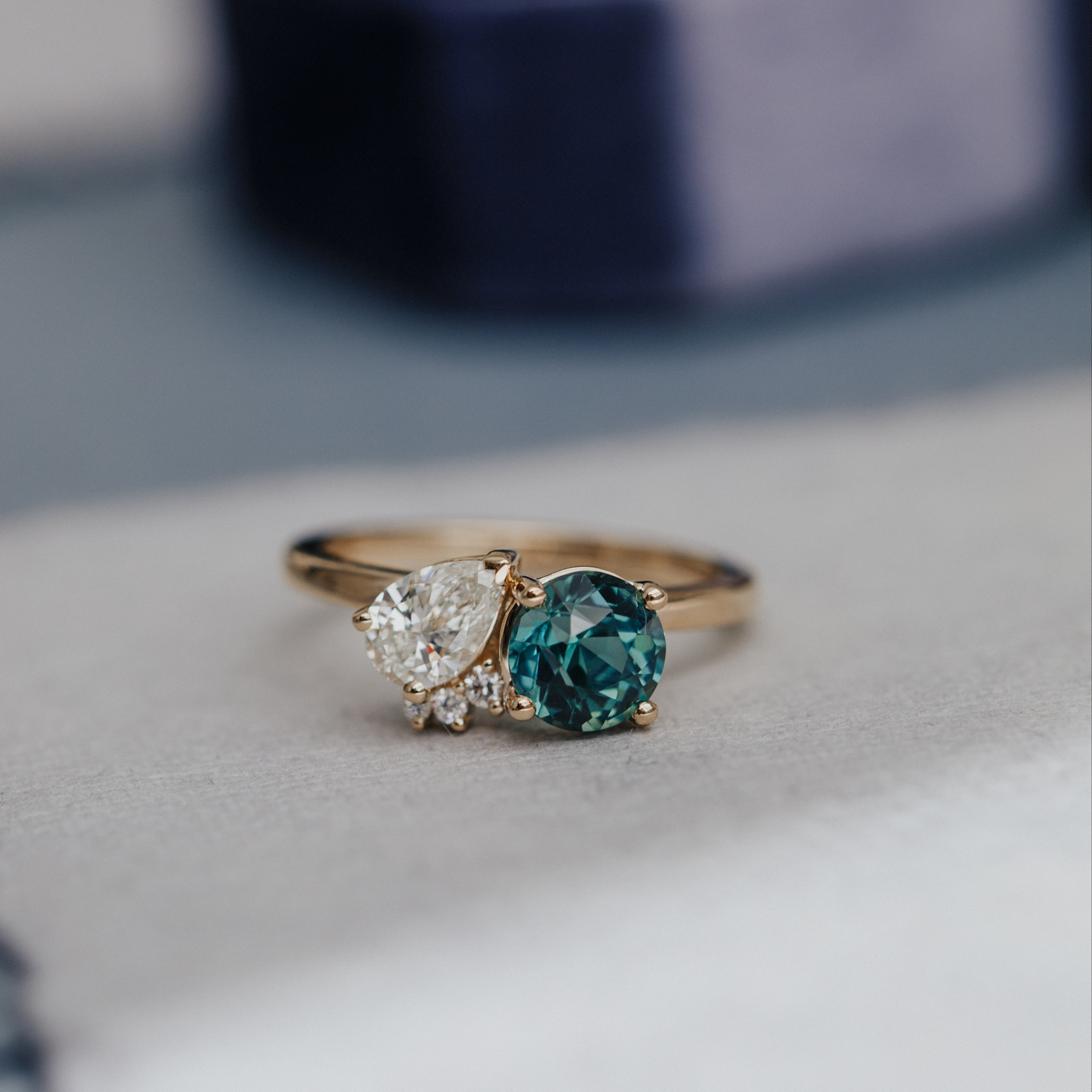The toi et moi round teal sapphire and pear diamond engagement ring on a neutral surface.