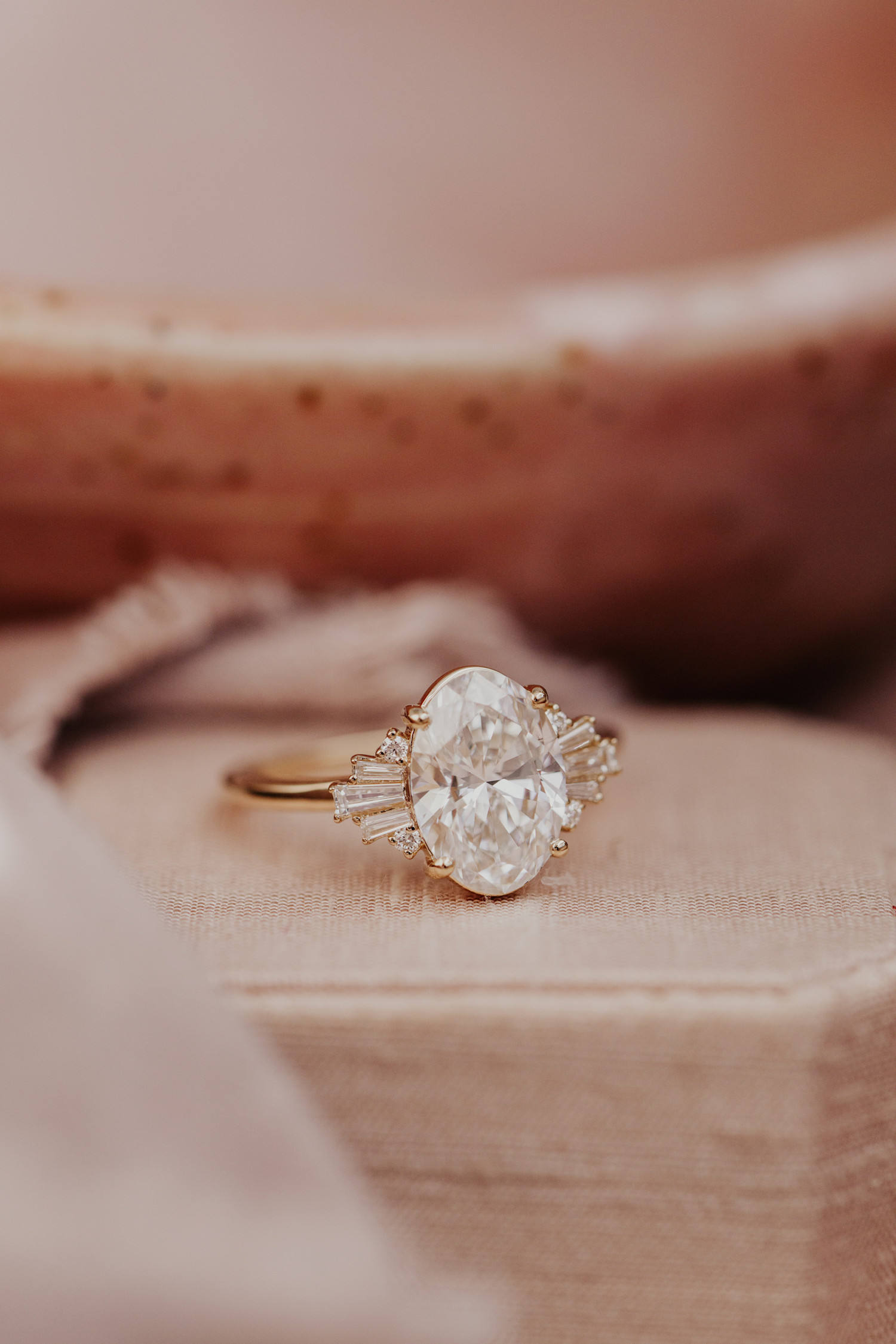 The Josephine oval diamond engagement ring on a pink textured surface.