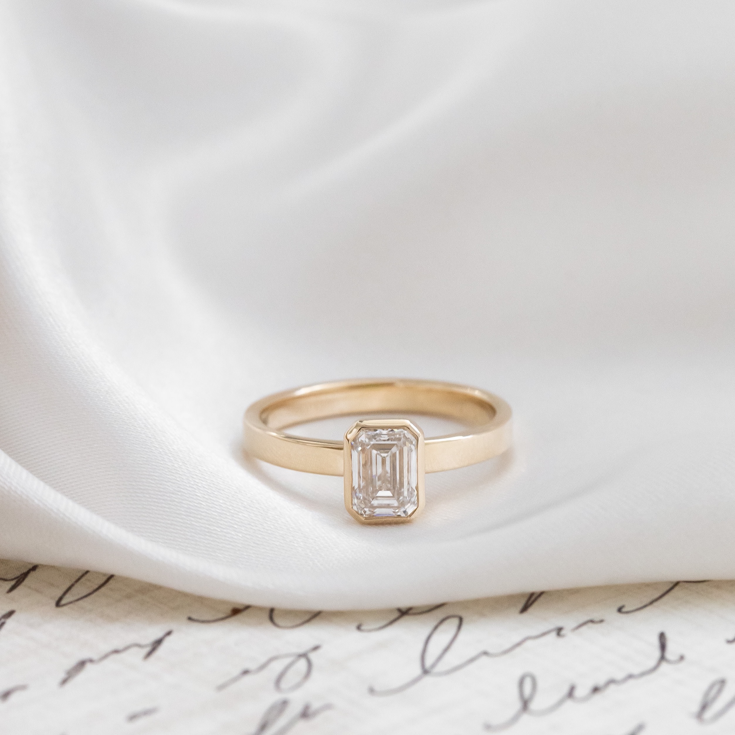 The Elena emerald cut solitaire diamond ring set in a yellow gold bezel and placed on a white silk fabric.
