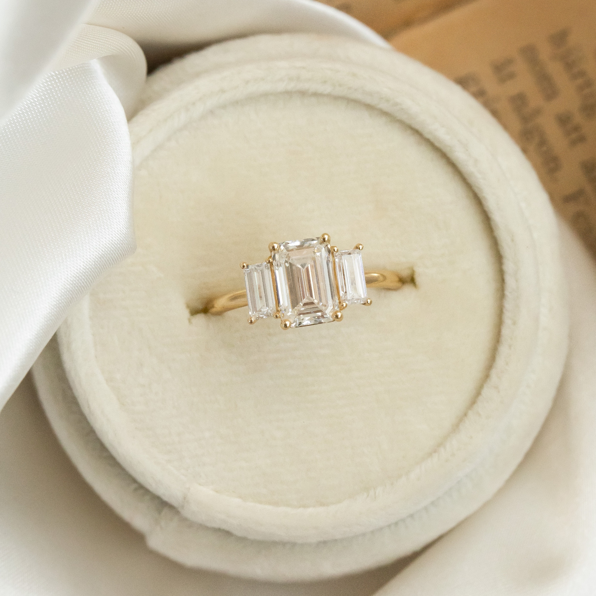 The Hannah emerald cut diamond engagement ring with diamond side stones placed in a white round ring box.