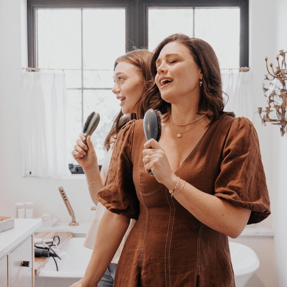 Mother and daughter signing into their hairbrushes. Mom is wearing stacked diamond necklaces.