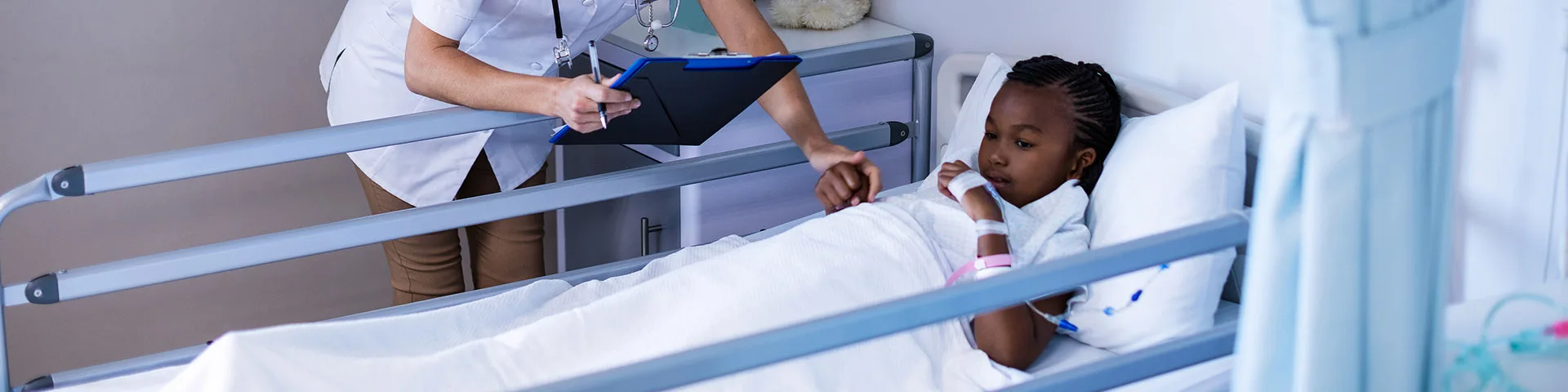 Adolescent patient patient laying in hospital bed accompanied by a smiling health care professional holding her hand. 