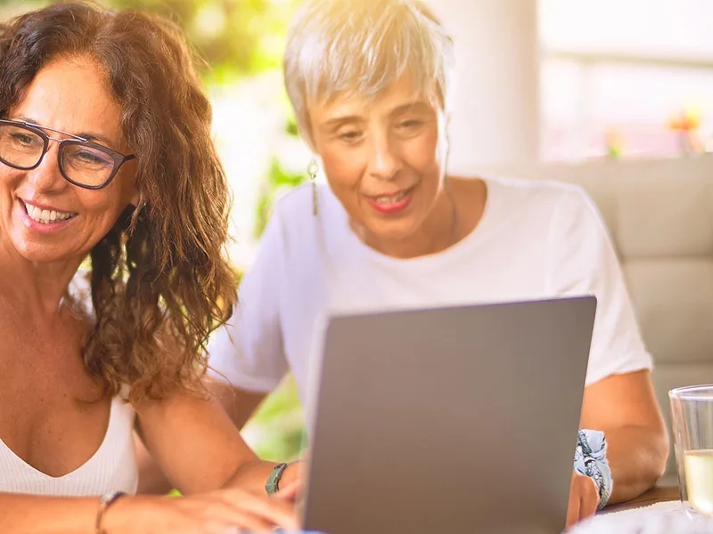 Two middle-aged women sitting at a table with a laptop and smiling.