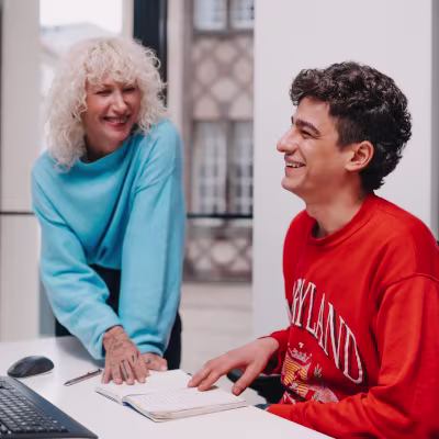 ATG Entertainment - Mann mit dunklen Haaren und rotem Oberteil am Schreibtisch mit Frau mit blauem Pullover und hellen Haaren