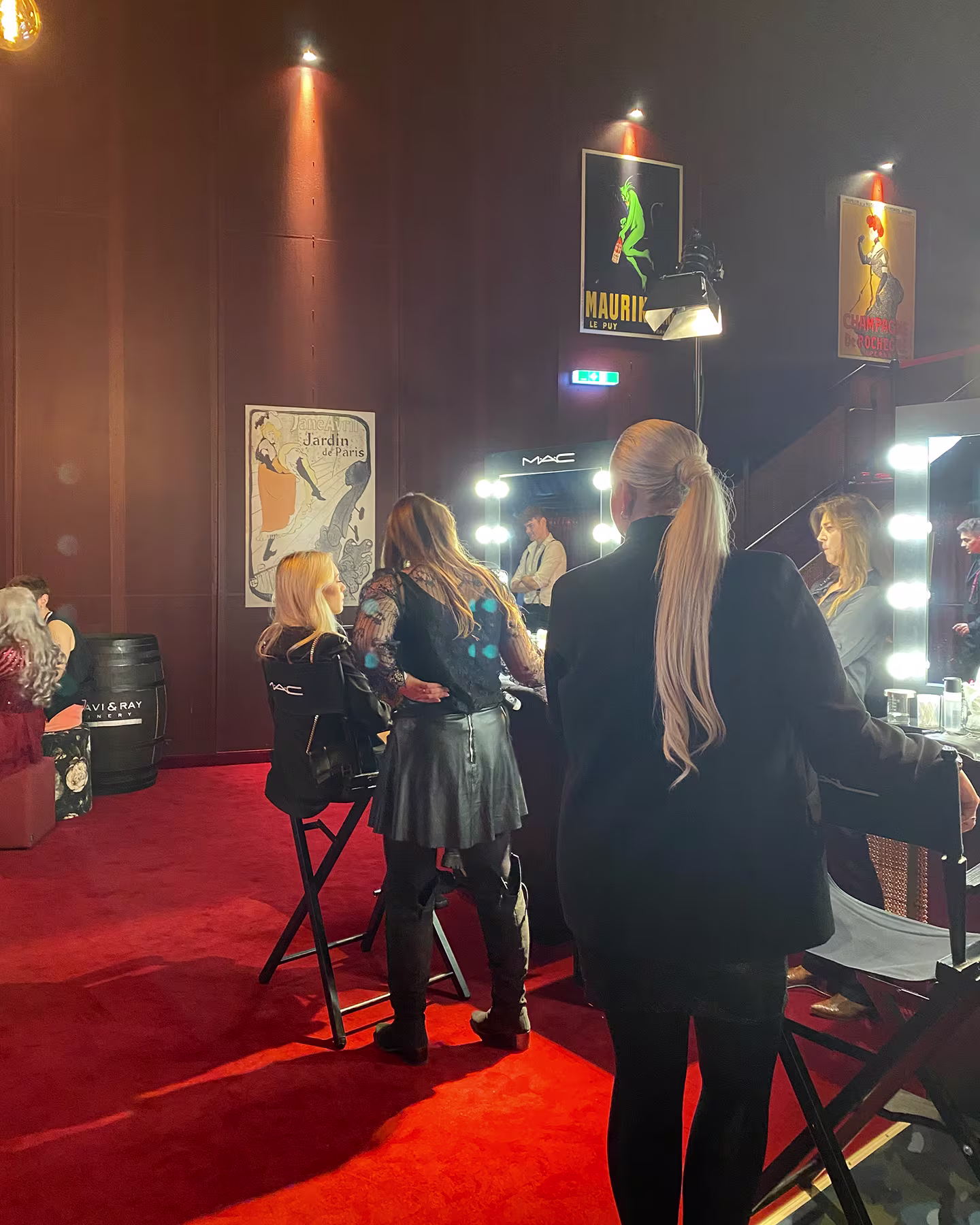 Backstage-Bereich mit beleuchteten MAC Schminkspiegeln und historischen Plakaten der Belle Époque, mehrere Gäste stehen auf rotem Teppich