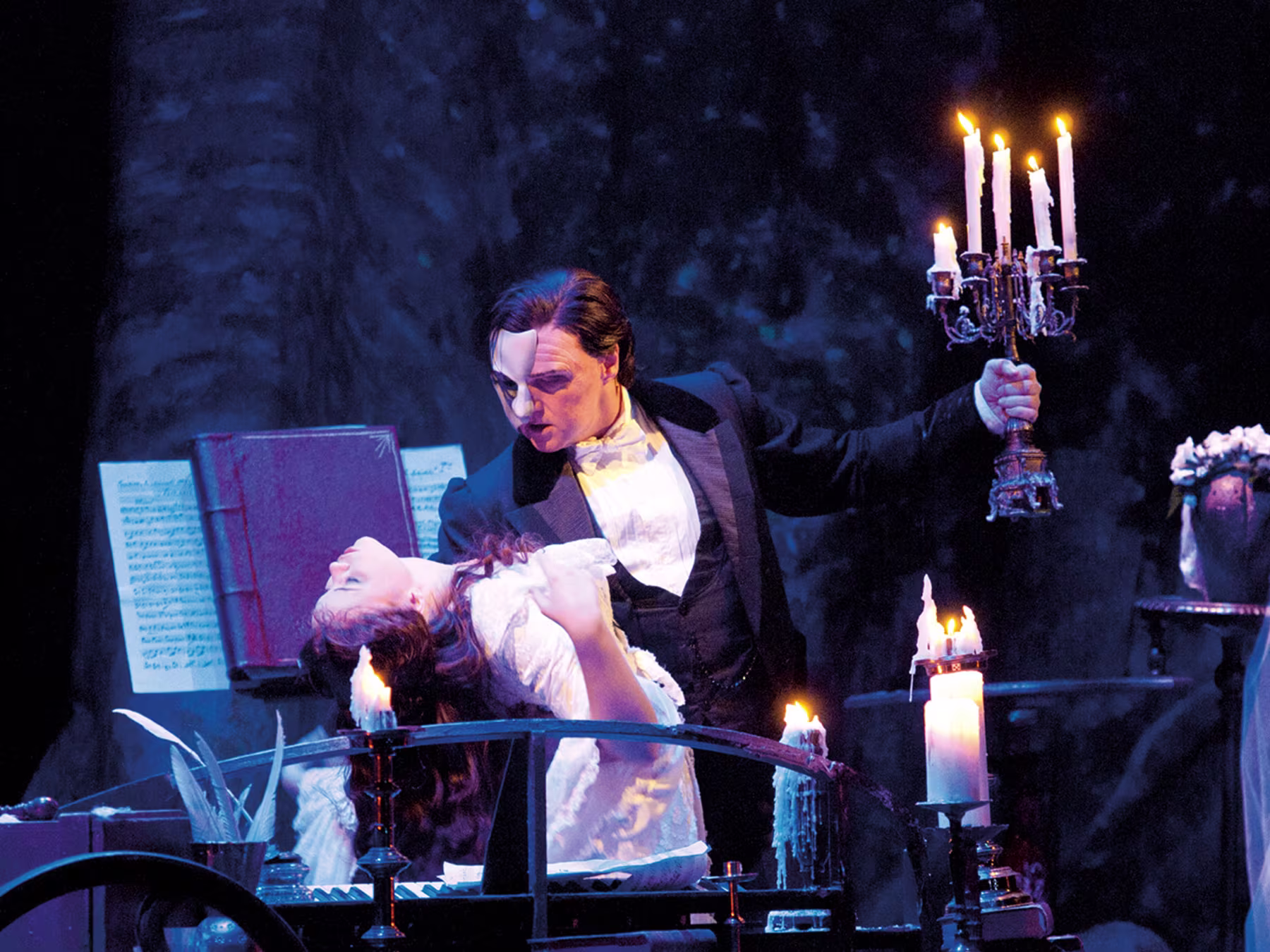 Das Phantom der Oper - Mann mit Maske hält Frau in weißem Kleid im Arm und in der anderen Hand hält er einen Kerzenständer mit brennenden Kerzen