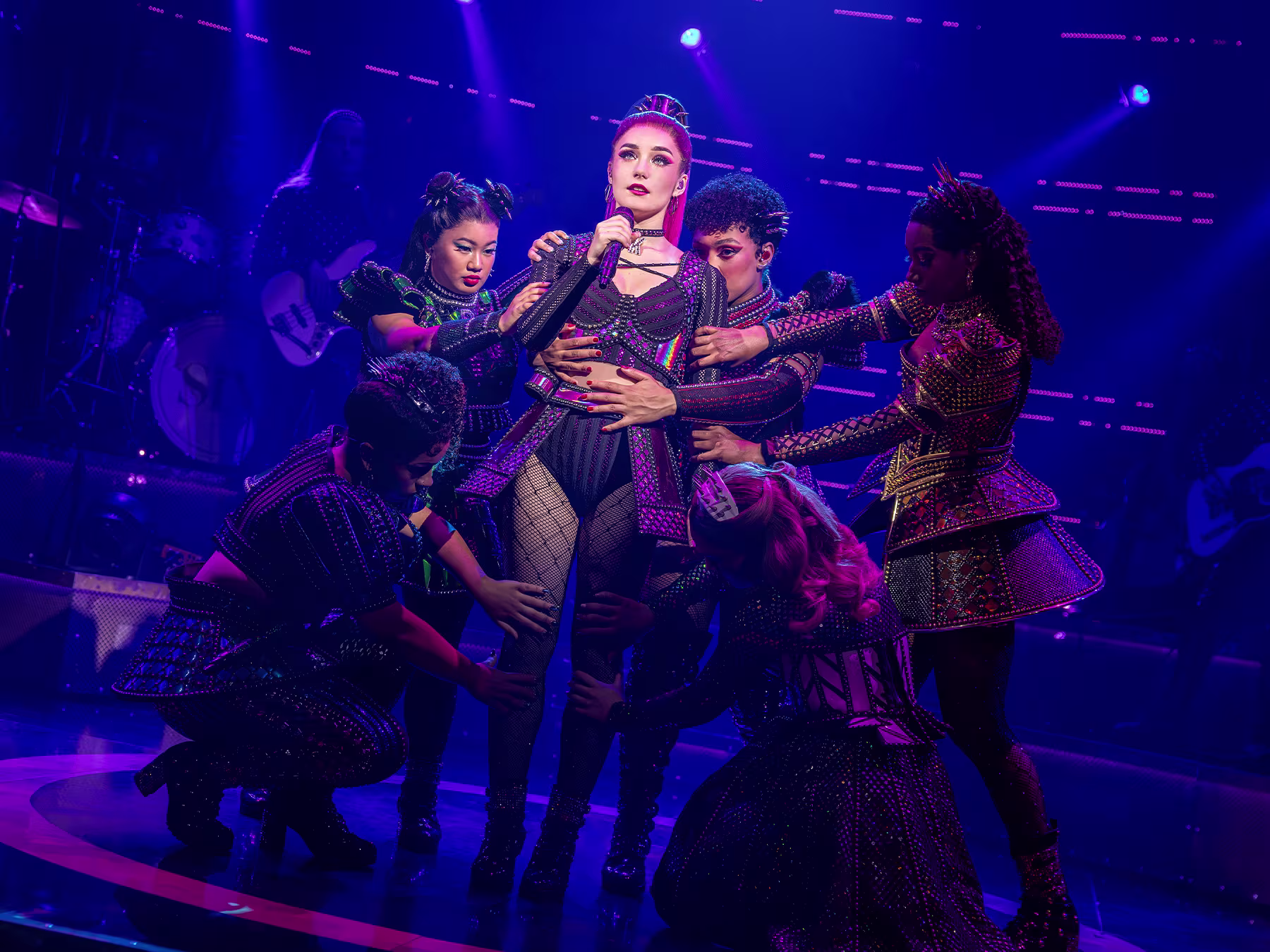 Foto aus der Show SIX: Königin steht auf der Bühne mit Mikrofon. Andere Fragen stehen um sie herum und berühren sie. 