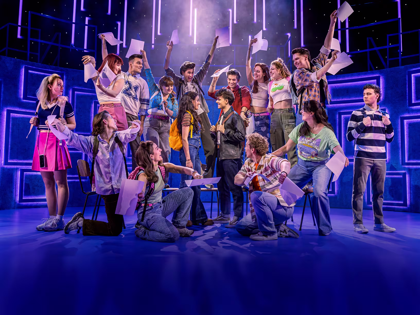 Eine Theater- oder Musicalszene mit einer Gruppe von Darstellern auf der Bühne. Sie halten Blätter in die Luft und befinden sich in verschiedenen dynamischen Posen, die Freude und Energie ausdrücken. Im Hintergrund ist ein schlichtes Bühnenbild mit blauen Streifen und Gebäudeelementen zu sehen. Die Darsteller tragen farbenfrohe Alltagskleidung, die die lebendige Stimmung der Szene unterstreicht.