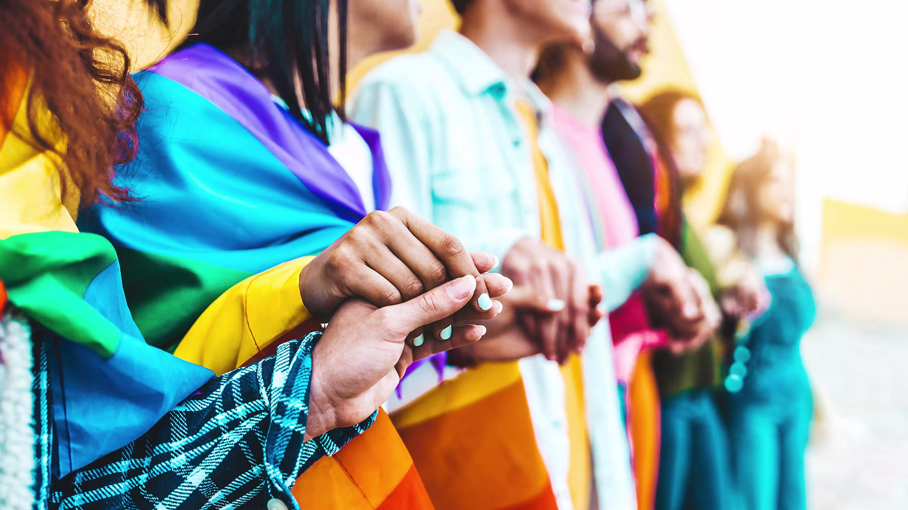 Mehrere Personen halten in Reihe ihre Hände. Sie tragen Regenbogenflaggen um ihre Schultern.