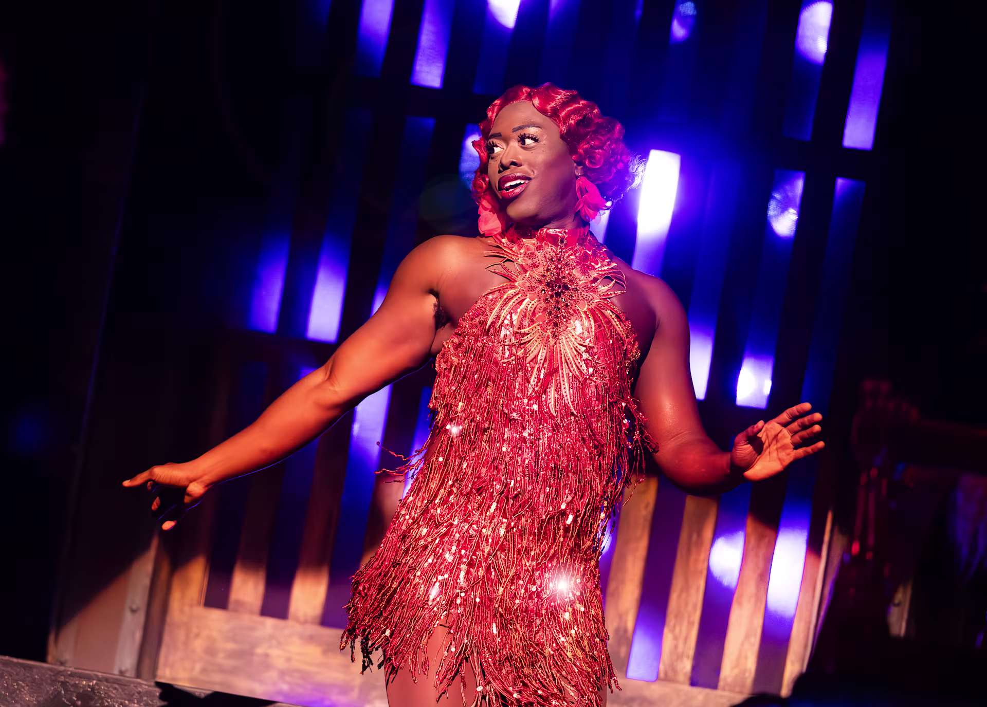 Kinky Boots – The Musical: Ein Darsteller in rotem Partykleid und mit roten Haaren lächelt das Publikum an.