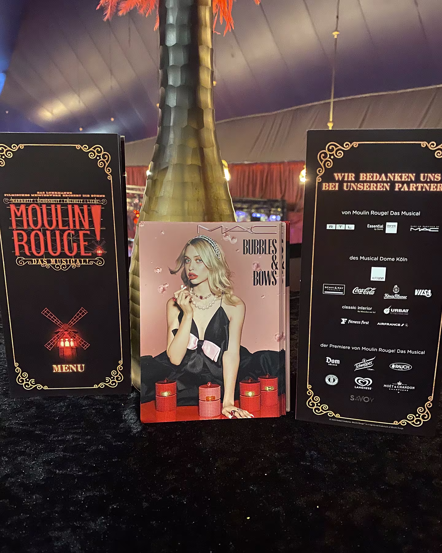 Menükarte von Moulin Rouge! Das Musical neben einer MAC Bubbles and Bows Broschüre und einer Partner-Danktafel vor einer goldenen Sektflasche