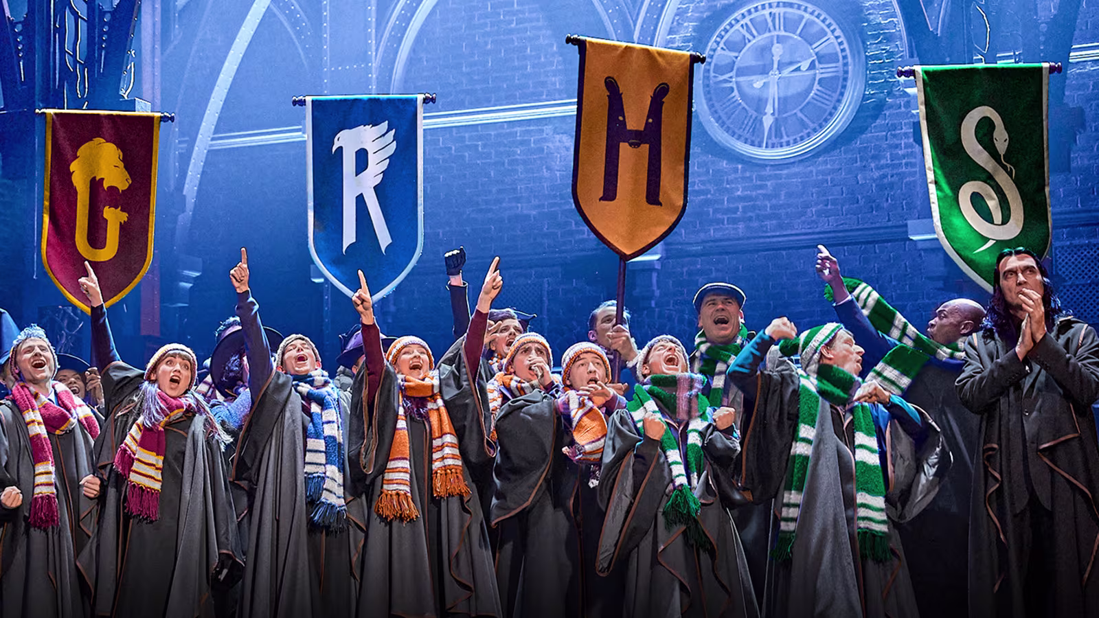 Eine jubelnde Gruppe in Hogwarts-Roben und Schals steht unter den Bannern der vier Häuser: Gryffindor, Ravenclaw, Hufflepuff und Slytherin. Im Hintergrund ist eine große Uhr sichtbar, die Szene stammt aus dem Harry Potter Musical.