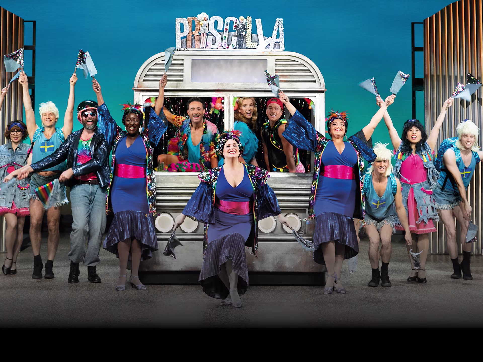 Szene aus “Priscilla – Queen of the Desert”: Drei Darsteller in auffälligen, glitzernden Kostümen performen auf einer bunten Bühne mit leuchtender Kulisse.