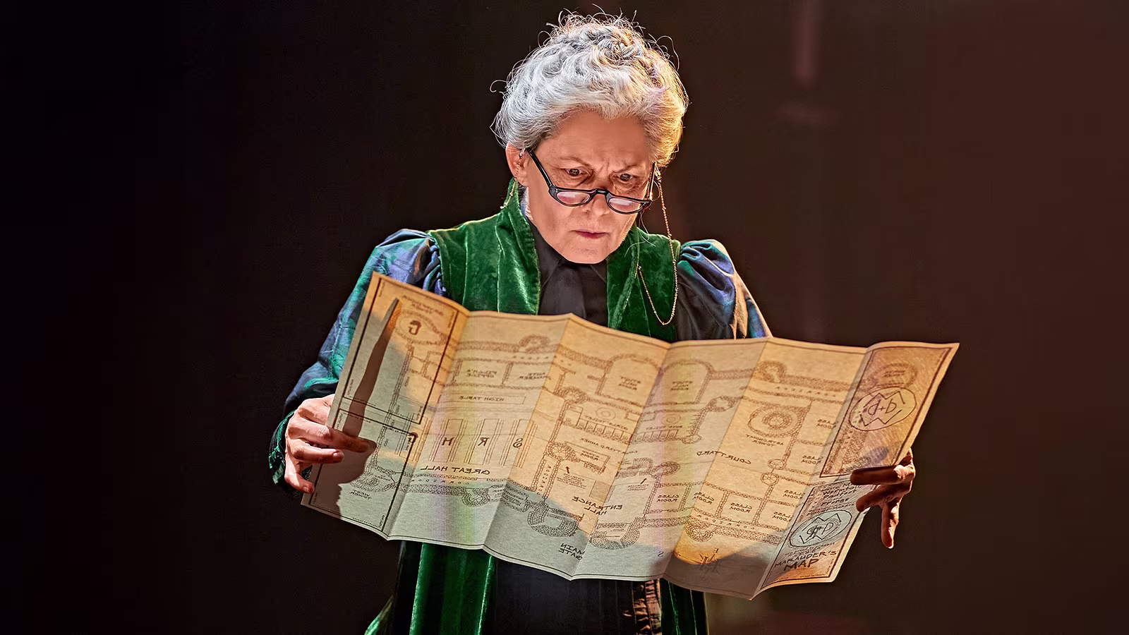 Das Bild zeigt eine ältere Frau mit Brille und grünem Umhang, die konzentriert eine detaillierte Karte studiert. Die Szene stammt aus dem Harry-Potter-Musical und vermittelt eine geheimnisvolle, fokussierte Atmosphäre.
