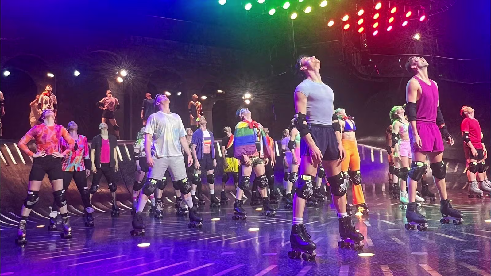 Tänzerinnen und Tänzer von Starlight Express in farbenfrohen Outfits und auf Rollschuhen stehen auf einer Bühne unter bunten, leuchtenden Scheinwerfern. Einige tragen Regenbogenkleidung, die auf eine Pride-Thematik hinweist, während alle nach oben schauen. Im Hintergrund sind weitere Tänzer und Rampen zu sehen, die Teil der Bühnengestaltung sind.