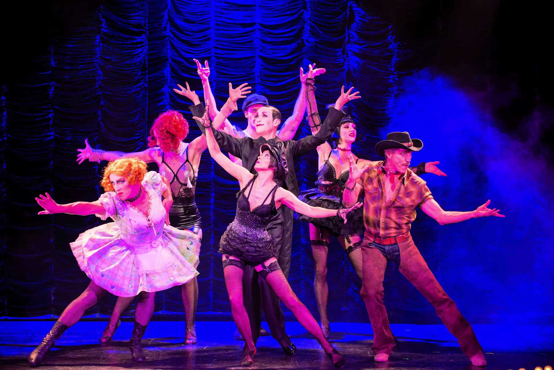 Szene aus einem Musical: Sieben Künstler*innen in extravaganten Kostümen, darunter Korsagen, Netzstrümpfe und Cowboy-Outfit, posieren ausdrucksstark vor einem blau-pink beleuchteten Vorhang.