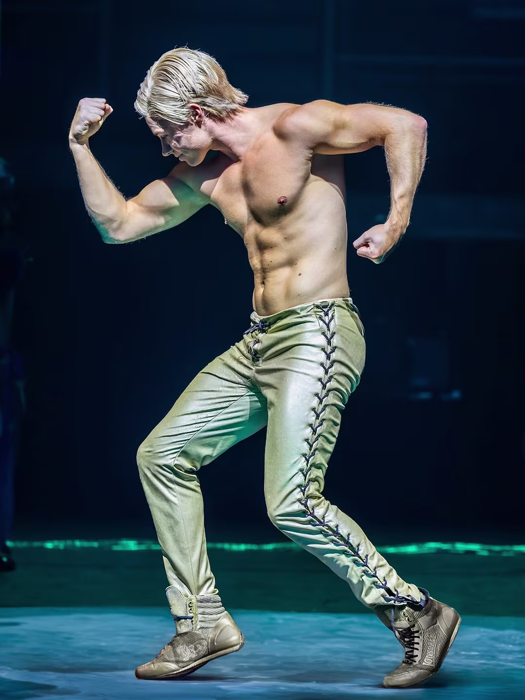 Szene aus der Rocky Horror Show: Ein muskulöser, oberkörperfreier Darsteller mit blondem Haar posiert in einer angespannten Haltung. Er trägt goldene, geschnürte Hosen und passende Schuhe. Der Hintergrund ist dunkel und die Bühne ist dezent beleuchtet.