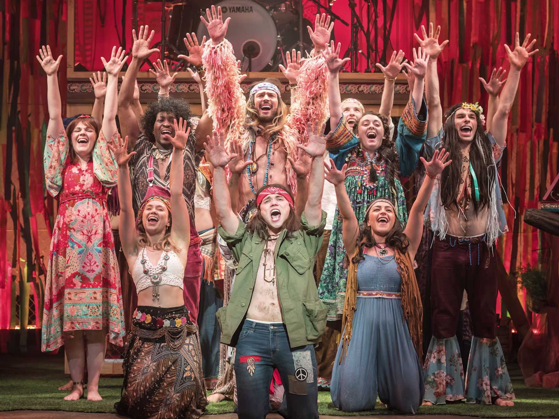 Szene aus “Hair – The Musical”: Eine Gruppe von Darstellern in bunten, hippie-inspirierten Outfits tanzt und singt auf einer farbenfrohen Bühne mit Blumenmotiven.