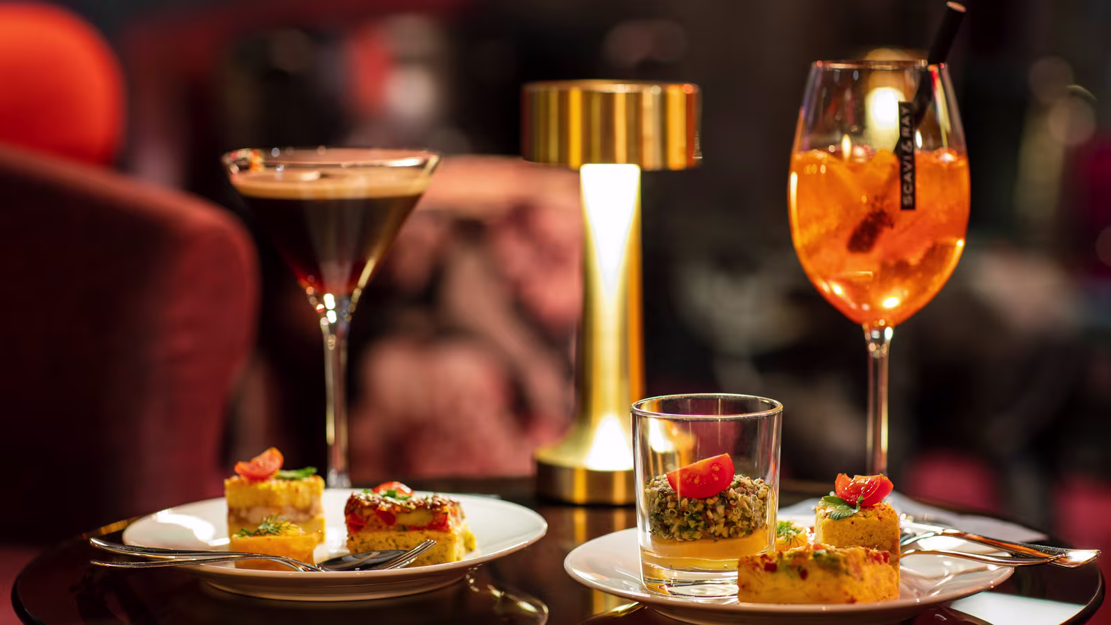 Ein stilvoll angerichteter Tisch mit Cocktails und kleinen Häppchen. Zu sehen sind ein Espresso-Martini, ein Aperol Spritz, sowie kunstvoll präsentierte Vorspeisen, begleitet von einer goldenen Tischleuchte, die für eine warme Atmosphäre sorgt.