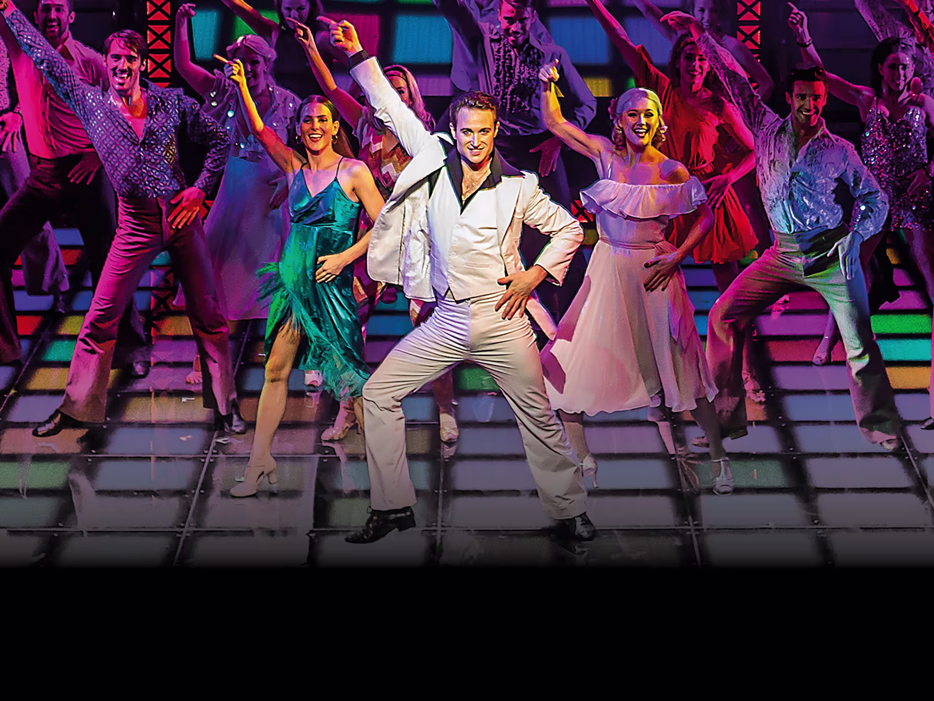 Eine Tanzszene aus “Saturday Night Fever” mit Tänzern in glamouröser Kleidung. Ein Mann in weißem Anzug steht im Mittelpunkt unter bunten Lichtern.