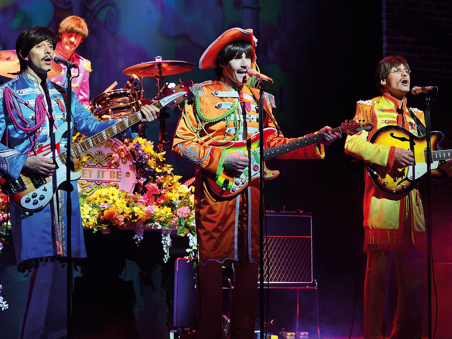 Szene aus “Let It Be”: Vier Musiker auf einer Bühne, die an die Beatles erinnern, spielen mit Retro-Instrumenten vor einer bunten Kulisse.