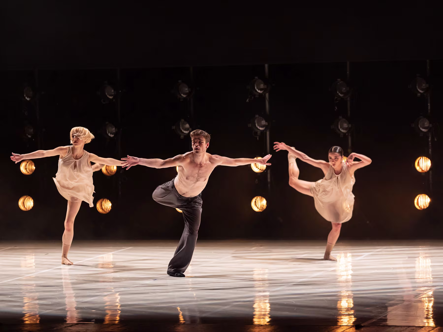 Drei Tänzer*innen tanzen im Spotlight auf einer dunklen Bühne. 