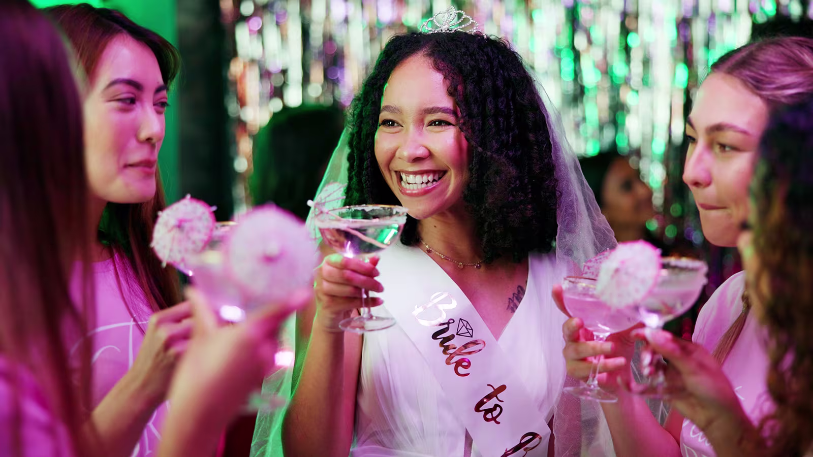 Braut mit Schleier und Schärpe feiert ihren Junggesellinnenabschied mit Freundinnen und Cocktails
