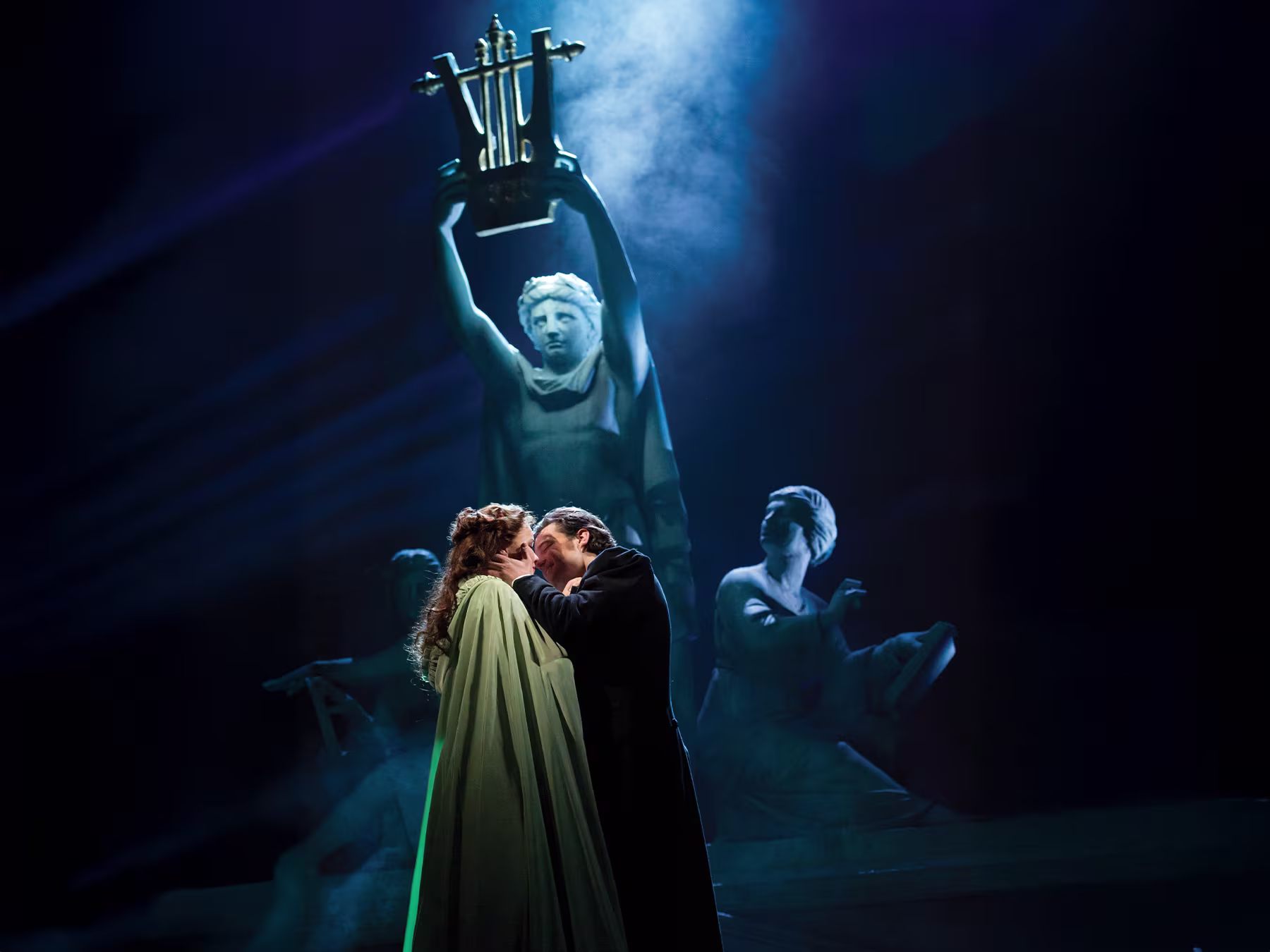 Das Phantom der Oper - Mann in dunklem Outfit und Frau mit grünem Mantel stehen im Dunkeln und küssen sich