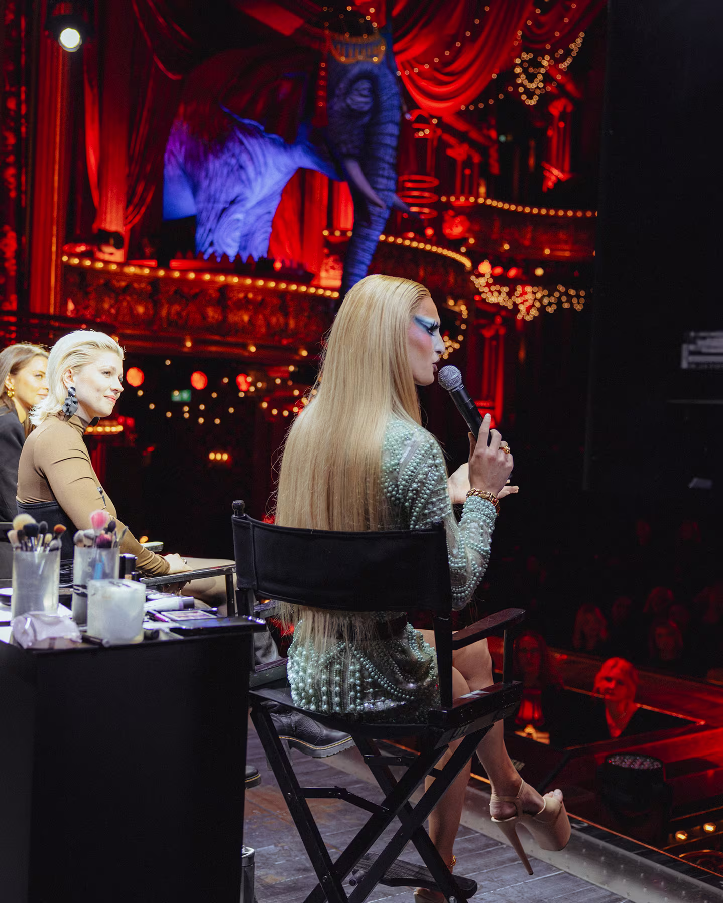 Zwei Frauen sitzen auf der Bühne von Moulin Rouge! Das Musical, eine Person mit langen blonden Haaren und Bühnen-Make-up spricht in ein Mikrofon, im Hintergrund der blaue Elefant