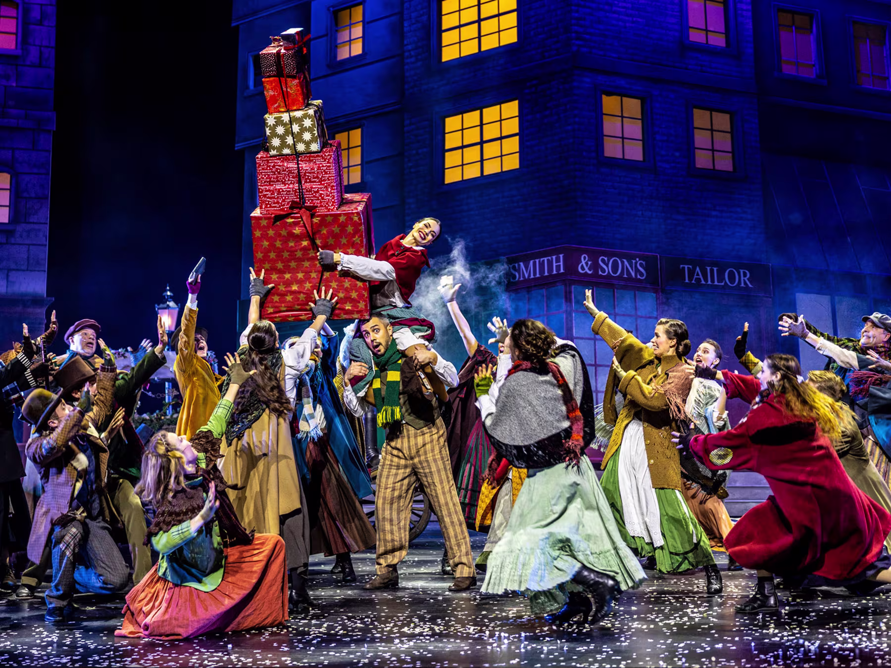 Der Geist der Weihnacht - Gruppe von Menschen mit bunten Kostümen auf einer Bühne. Person in der Mitte trägt viele Geschenke. 