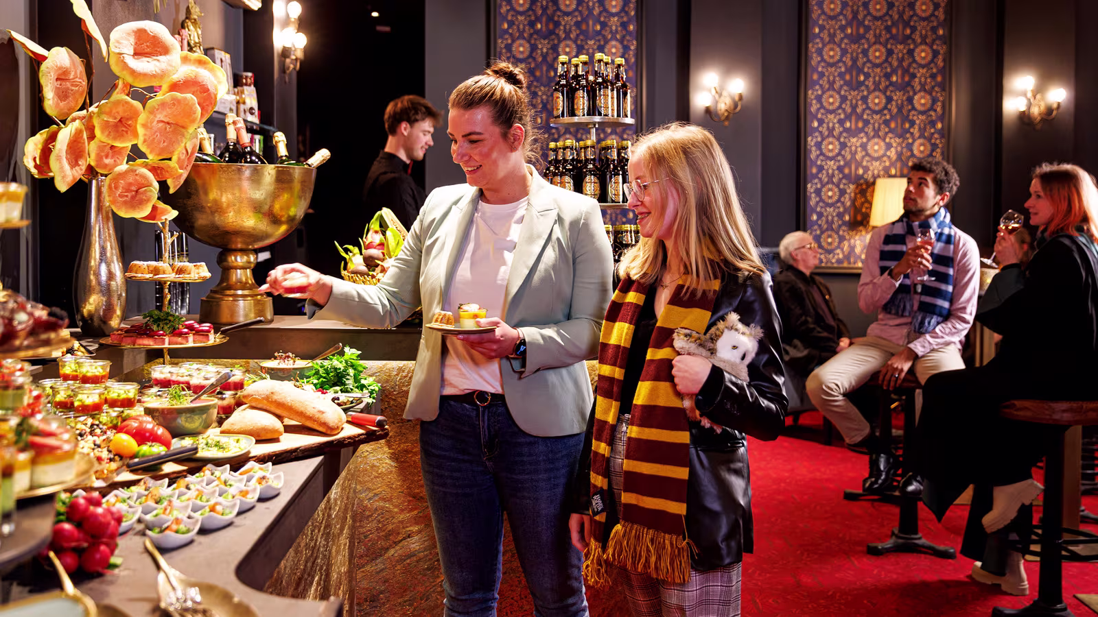 Zwei Frauen stehen an einem dekorativen Buffet, eine trägt einen rot-gelben Schal. Im eleganten Lounge-Hintergrund genießen weitere Personen Getränke.