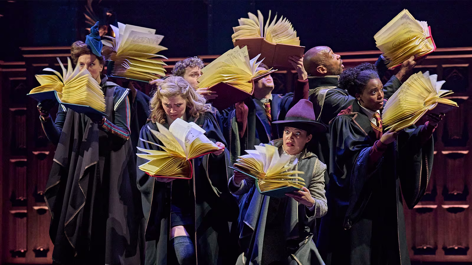 Das Bild zeigt eine Szene aus dem Harry-Potter-Musical, in der mehrere Figuren in Zauberumhängen lebhaft mit magischen Büchern interagieren. Die Bücher scheinen sich selbstständig zu bewegen, während die Darsteller konzentriert und erstaunt wirken.