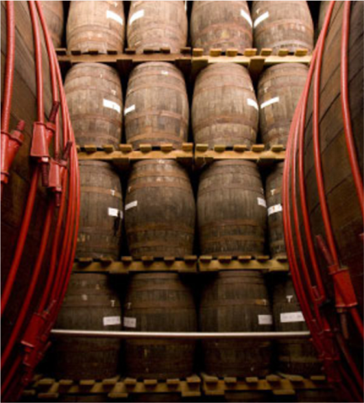 A collection of stacked wooden whisky barrels in a warehouse, surrounded by large vats with red metal hoops.