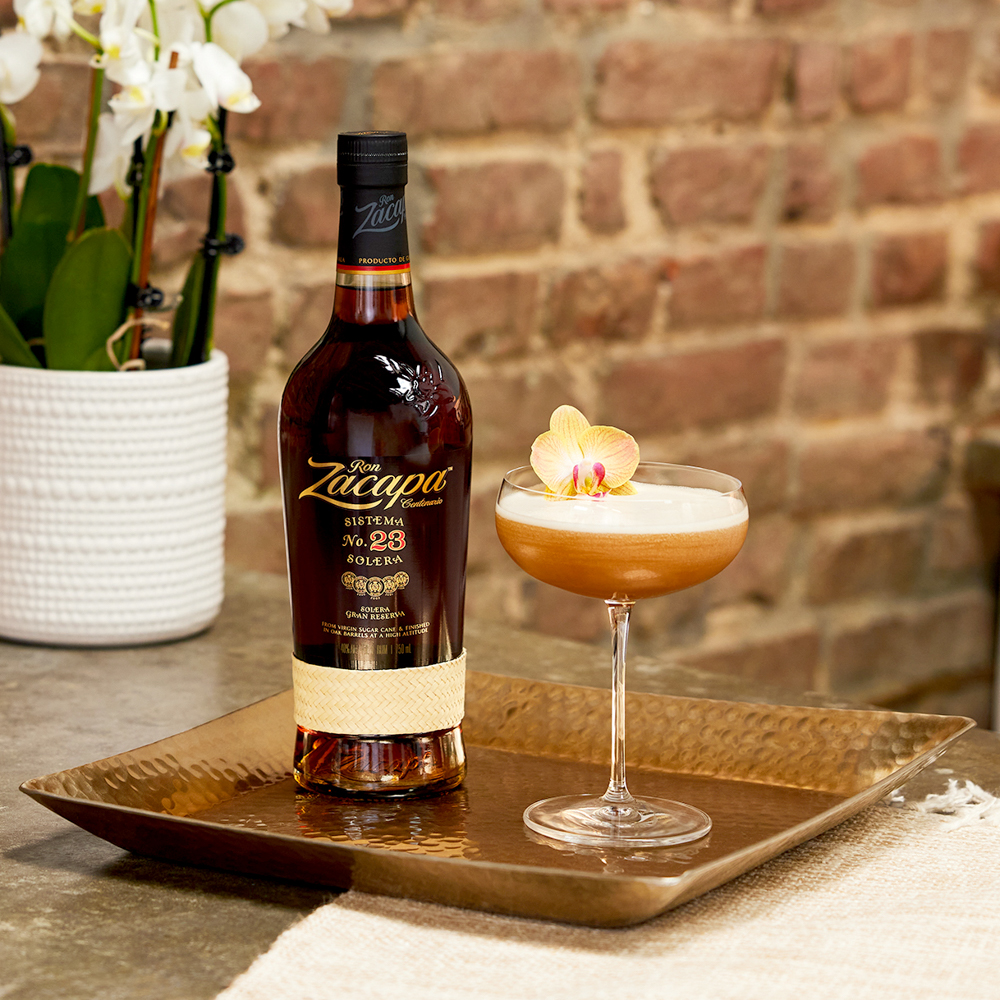 A bottle of Ron Zacapa Sistema 23 Solera rum is placed on a textured, gold-toned tray beside a coupe glass filled with a frothy cocktail.