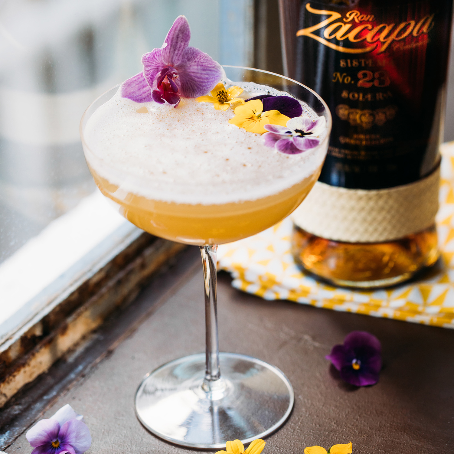 A cocktail glass filled with a frothy Ron Zacapa rum drink, garnished with vibrant purple and yellow edible flowers.