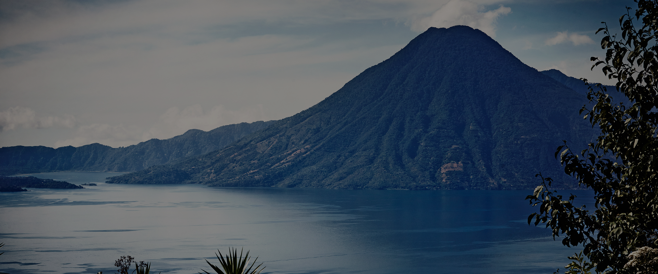 A majestic mountain rises from the edge of a calm lake, surrounded by smaller hills under a cloudy sky.