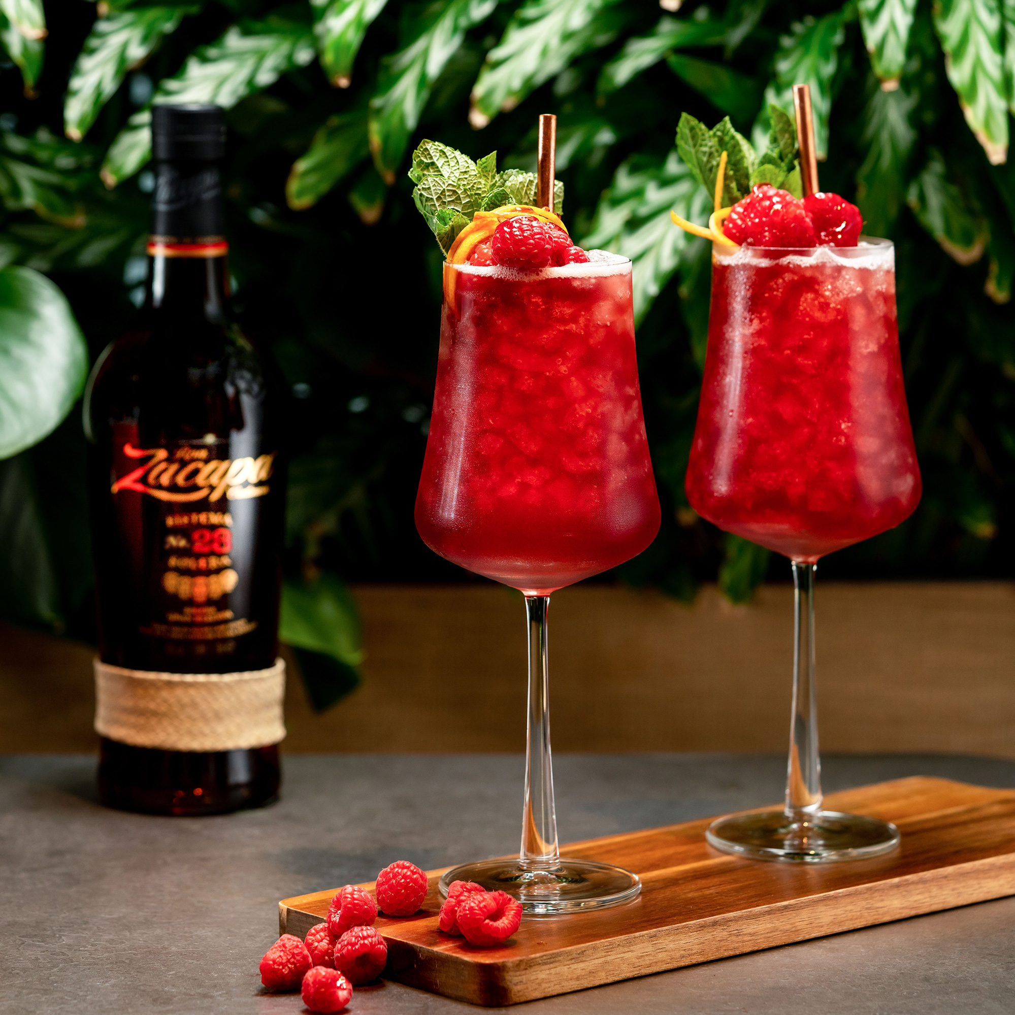 Two elegant stemmed glasses filled with a vibrant red cocktail, topped with crushed ice and garnished with fresh raspberries, mint leaves, and orange peel.